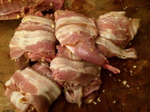 Season the marinated rabbit all over with salt and pepper, then wrap it with the bacon strips and tie them into place. This is a bit of an athletic endeavor: be calm and go slowly.  (I lay the bacon strips on the cutting board, slightly overlapping at the ends. I place the rabbit atop them, then slide the string under the bacon and bring it up – and the bacon with it – and around the rabbit, tying firmly to hold the bacon in place.) Use as many pieces of string as you need. If some of the bacon refuses to get caught in the string, simply skewer them into the rabbit using trussing skewers.